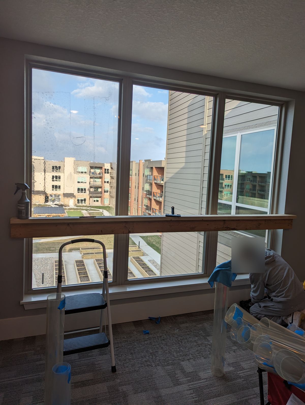 Installer applying clear security window film to a large three-pane apartment window in Sioux Falls, with film rolls and a step stool in the foreground.